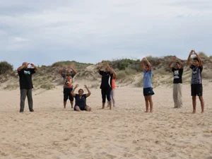 A group practising Wayapa Wuurrk on a sandy beach, arms raised in a shared movement and wellbeing session.