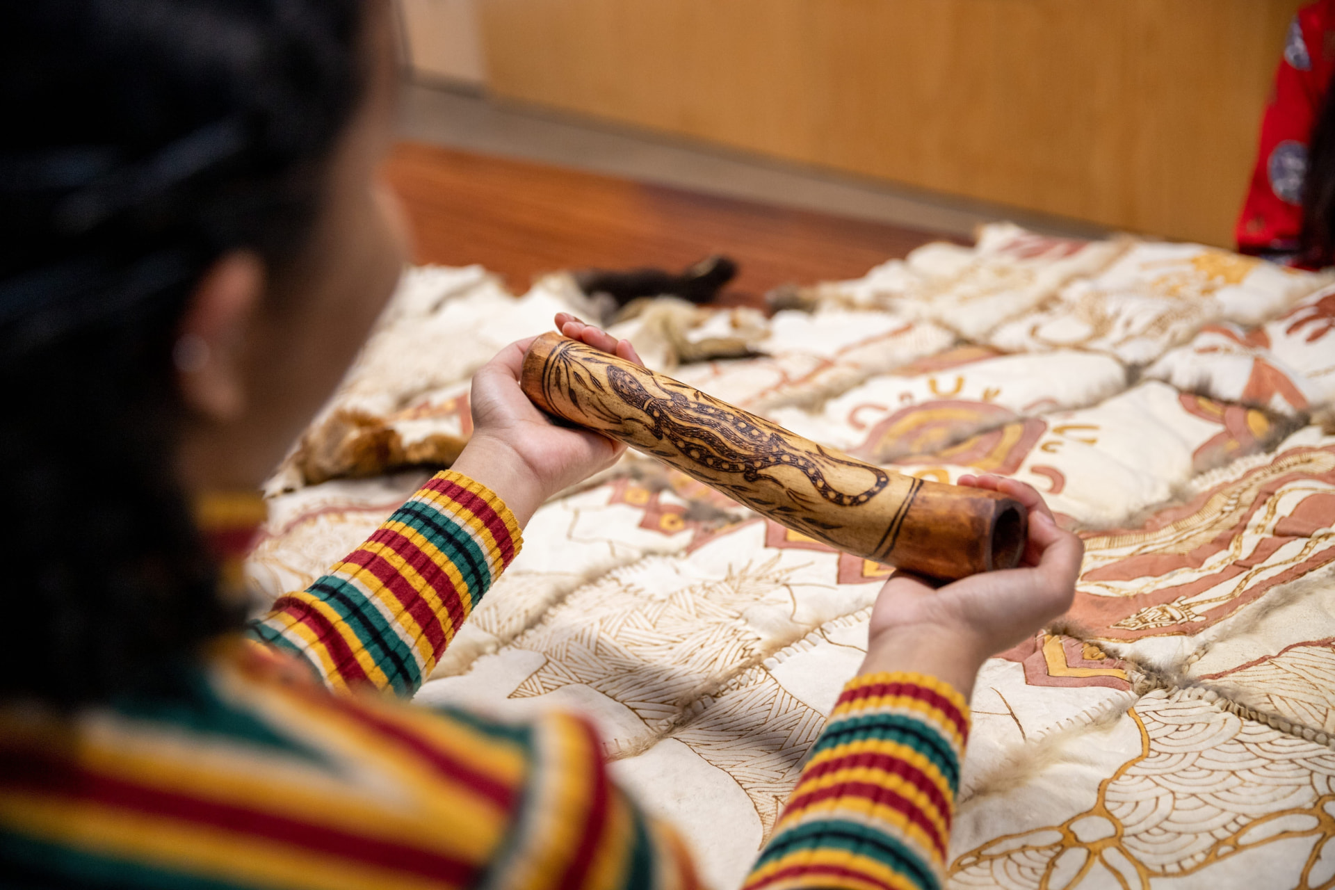 A carved wooden cultural object with painted designs, held by a First Nations practitioner in a colourful woven garment.