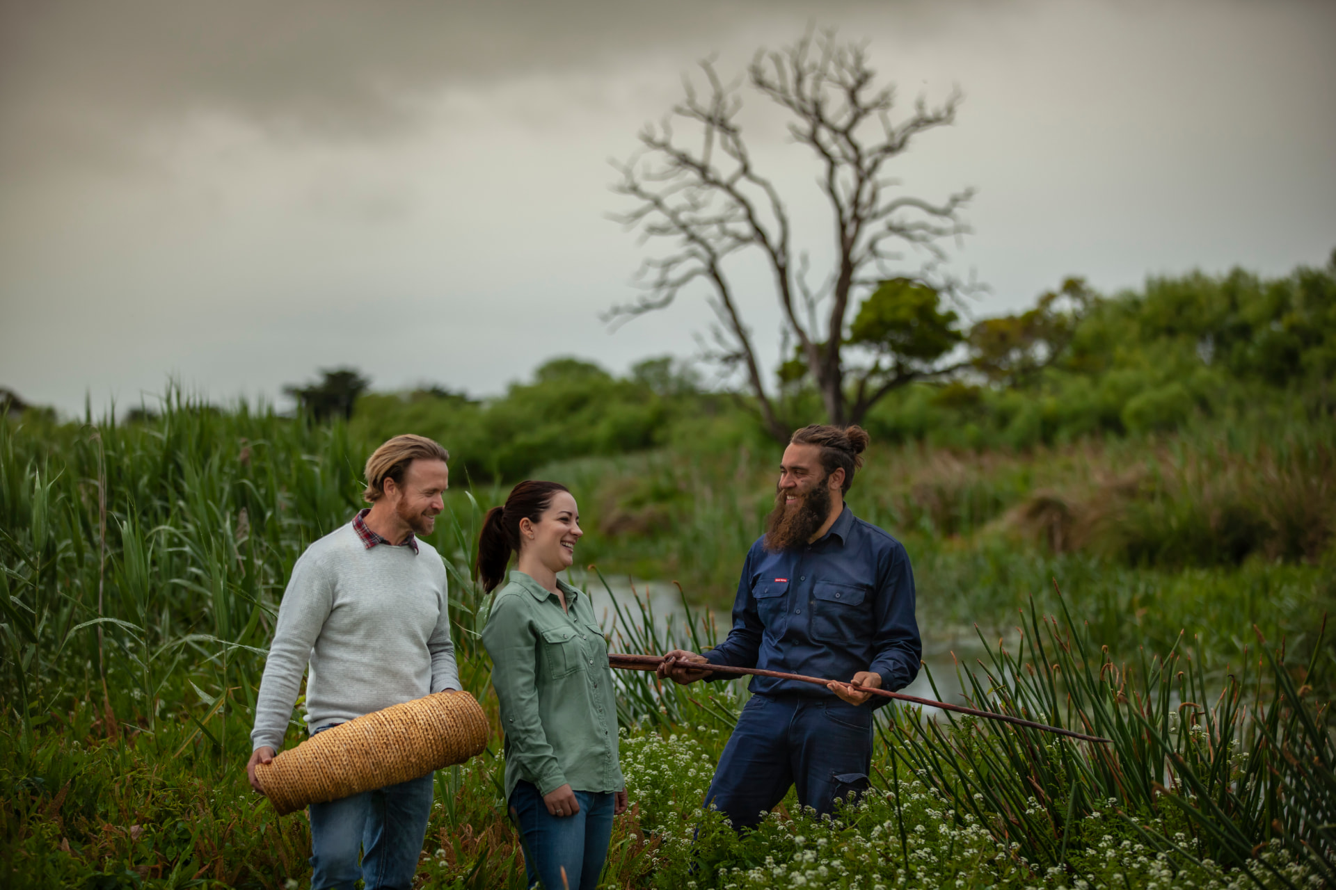 Three people on Country in regional Victoria, one carrying a woven mat and another holding a wooden pole, with wetlands and old gums behind them.