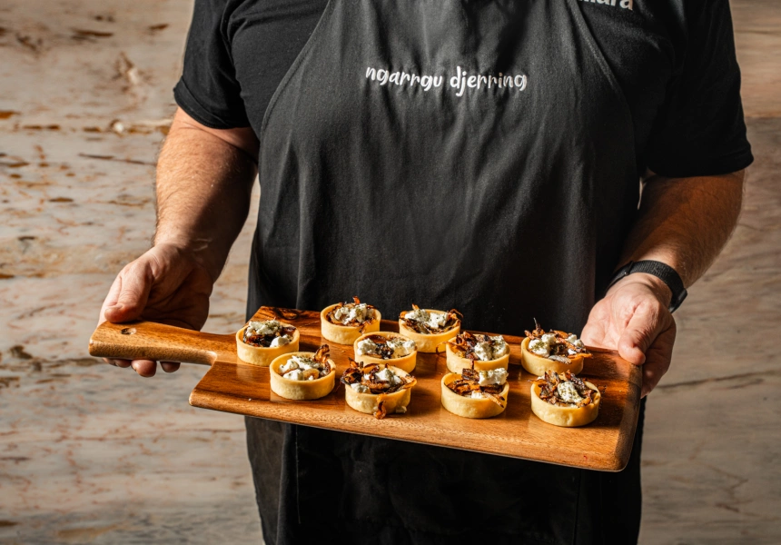 A chef holding a wooden board of small savoury tarts topped with native ingredients.
