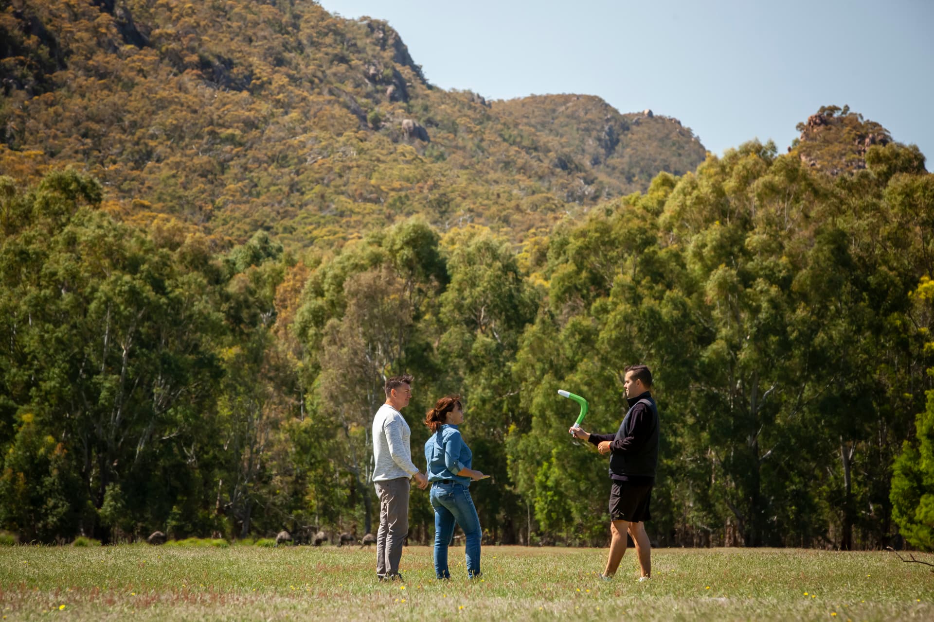 A First Nations guide demonstrating a boomerang to visitors on open Country with mountains behind.
