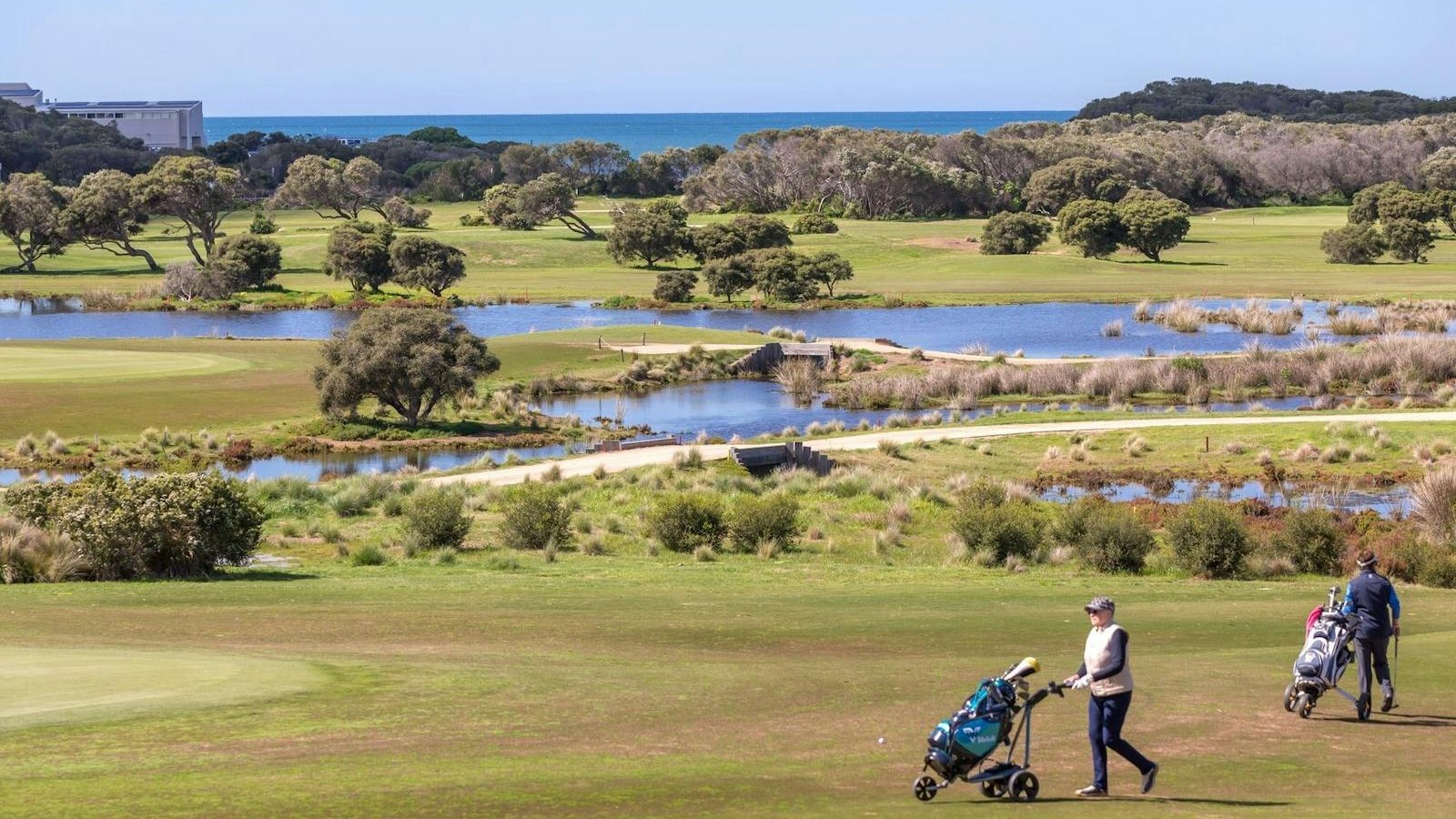 Golfers walking a fairway at RACV Torquay with wetlands, coastal scrub and Bass Strait behind